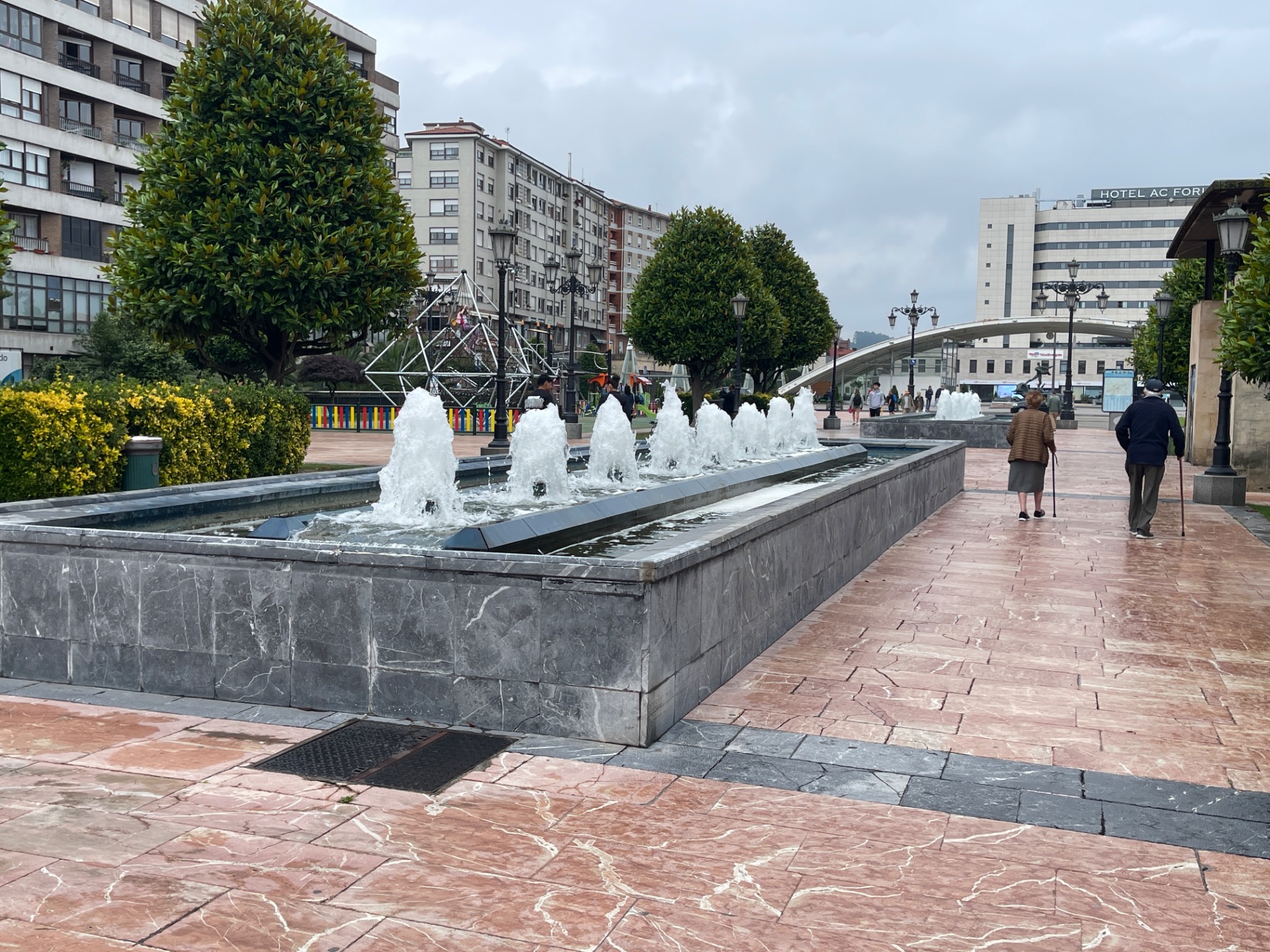 Fuente con chorros de agua en plaza, árboles y edificios alrededor, personas caminando bajo cielo nublado.